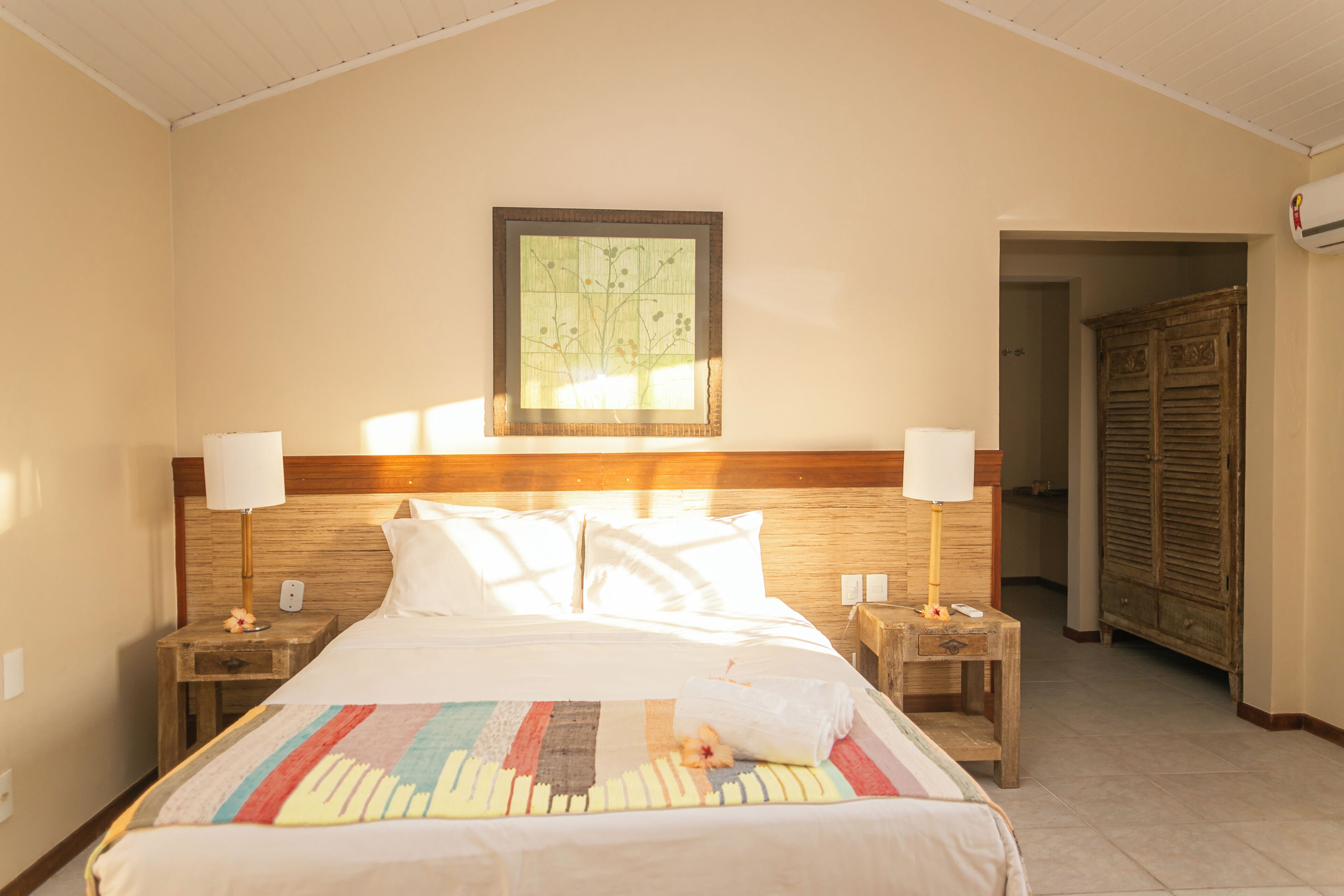 A hotel room with a large bed, wooden headboard, two bedside tables with lamps, a wall-mounted air conditioner, and a door leading to another room.
