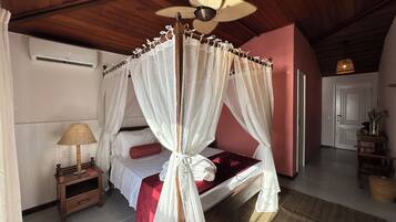 A bedroom with a four-poster bed, a ceiling fan, a bedside table with a lamp, and a red accent pillow.
