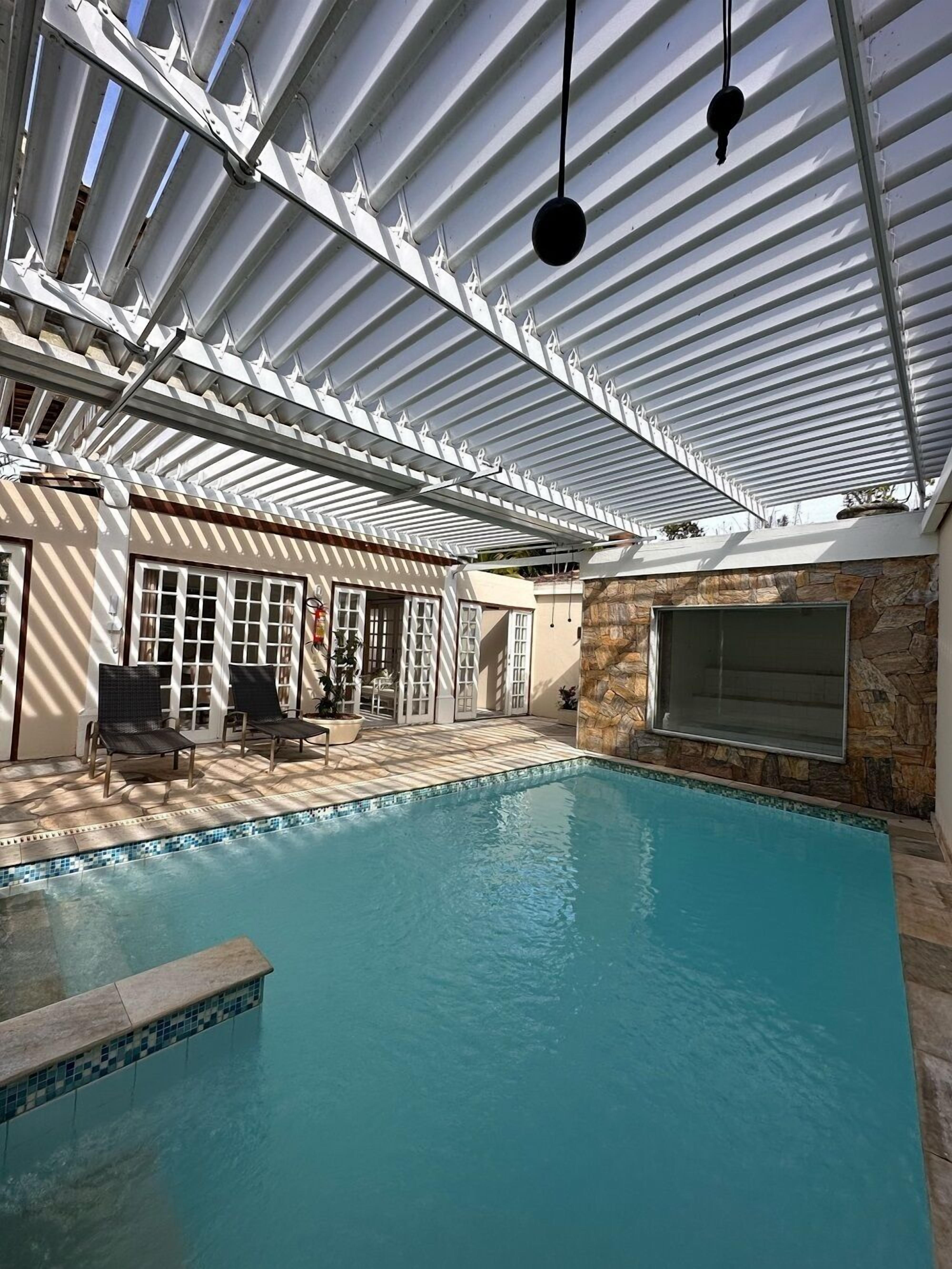 Una piscina interior con borde de azulejos, pared de piedra y zona de patio techada con sillas y una puerta.