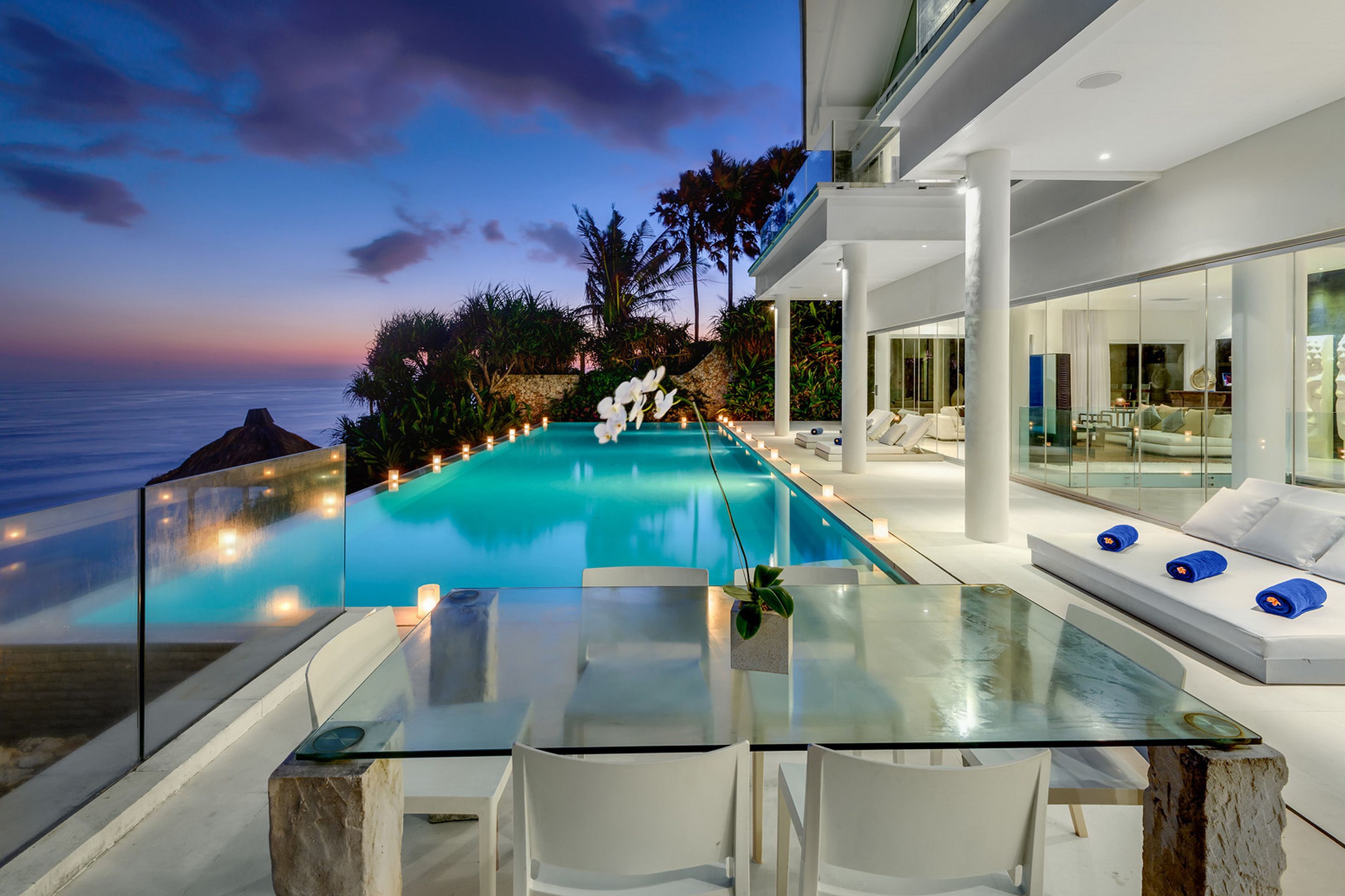 A modern infinity pool with a glass edge, white furniture, and a view of the ocean at dusk.