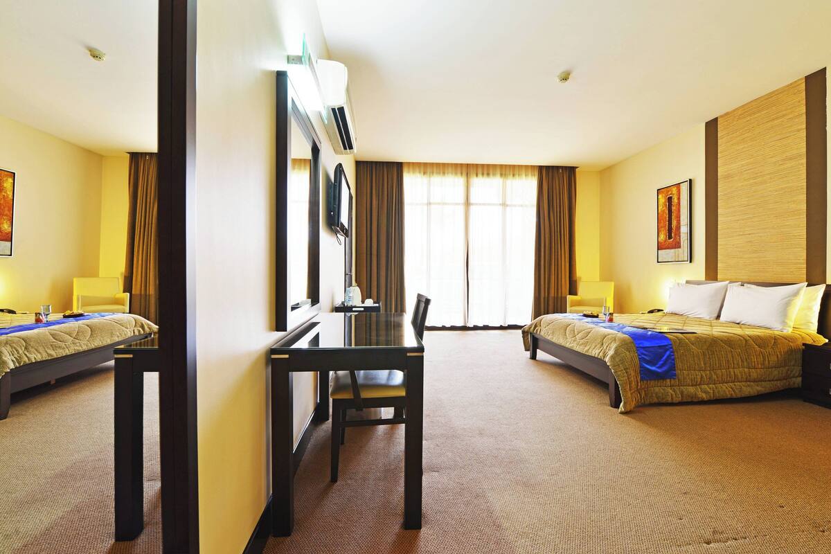 Habitación de hotel con dos camas, un escritorio, una silla, un televisor y un ventanal grande con cortinas.