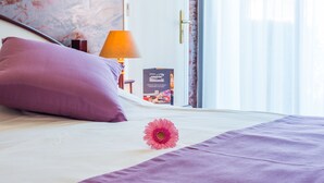 Un lit bien fait, avec un oreiller violet et une fleur rose, une lampe et un tableau encadré accroché au mur.