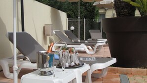 Un espace extérieur aménagé pour les repas, avec des tables et des chaises blanches, un grand bac à fleurs et des parasols pour faire de l’ombre.