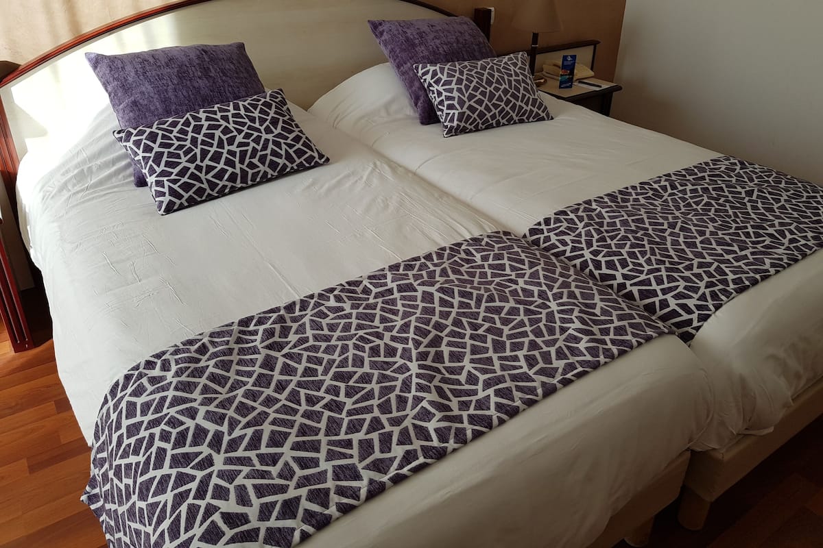 A double bed with a white and patterned bedspread, two purple pillows, and a nightstand with a lamp and a phone.