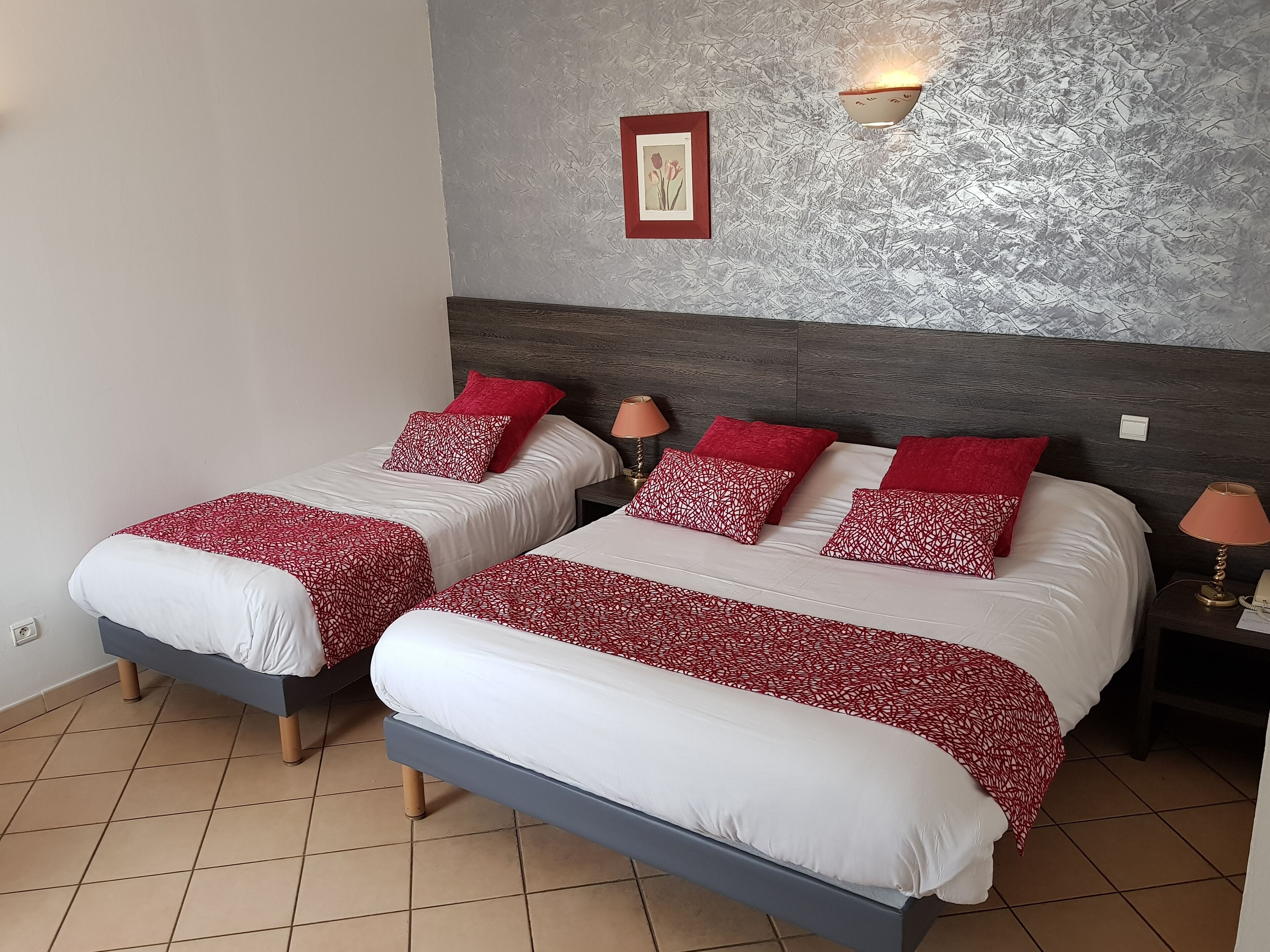 A hotel room with two beds, each with white and red patterned bedding, a grey headboard, a wall-mounted lamp, and a framed picture.
