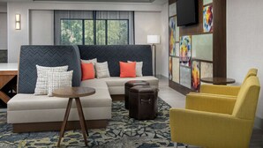 Une chambre d’hôtel moderne avec un canapé d’angle, un fauteuil jaune, une table ronde en bois et une grande fenêtre donnant sur la forêt.