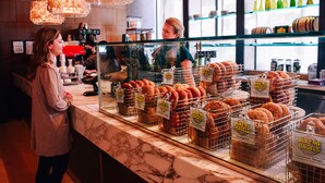Mostrador de panadería con varios bagels y productos de pastelería expuestos en cestas de alambre, una máquina de café y una caja registradora.