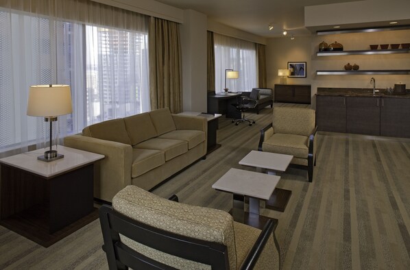 A hotel room with a sofa, armchair, small tables, a desk, and a kitchenette.