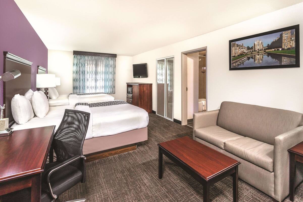 Habitación de hotel con cama, sofá, escritorio, silla, televisor y un cuadro en la pared.