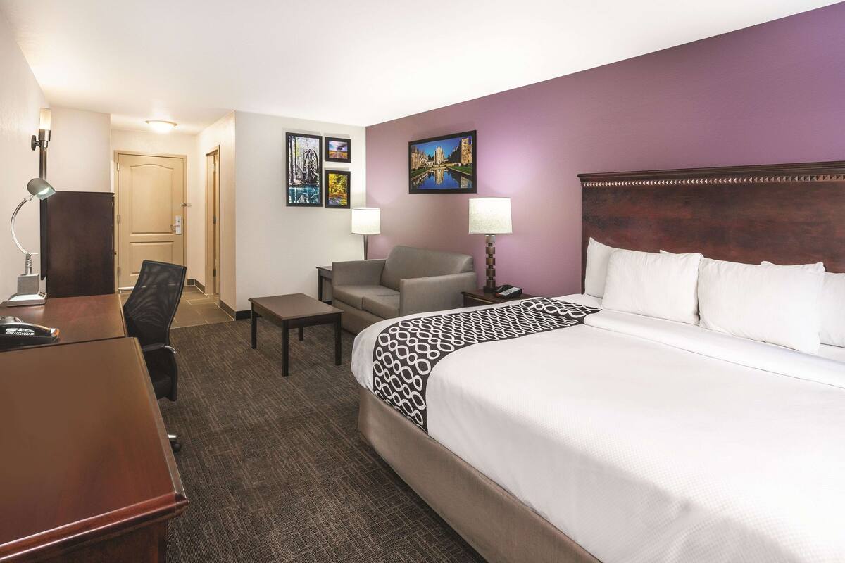 Habitación de hotel con una cama grande, un escritorio, una silla y dos l ámparas. Hay cuadros enmarcados en la pared y una puerta al fondo.