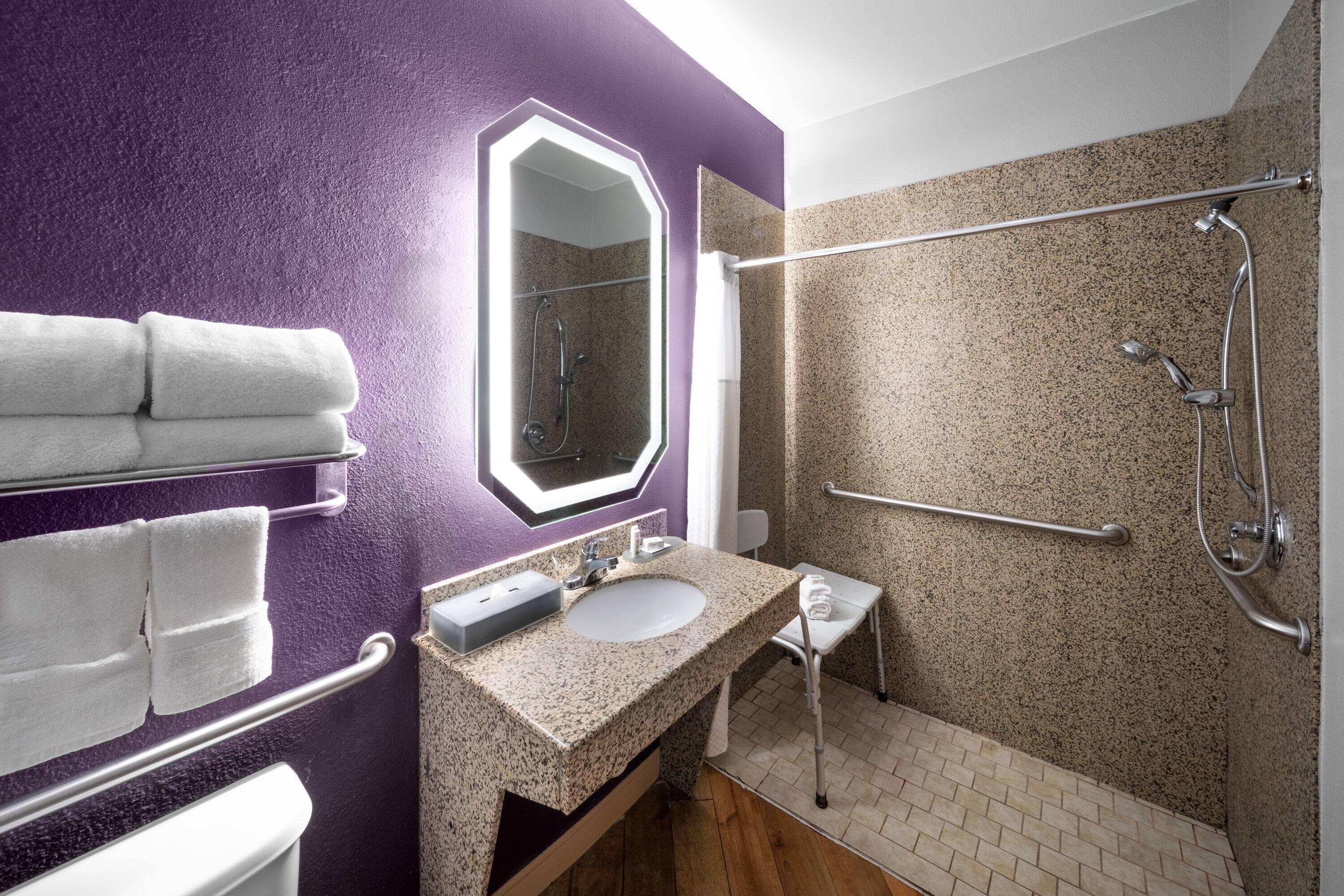 A bathroom with a purple wall, a sink with a mirror, a shower with a grab bar, and towels on a rack.