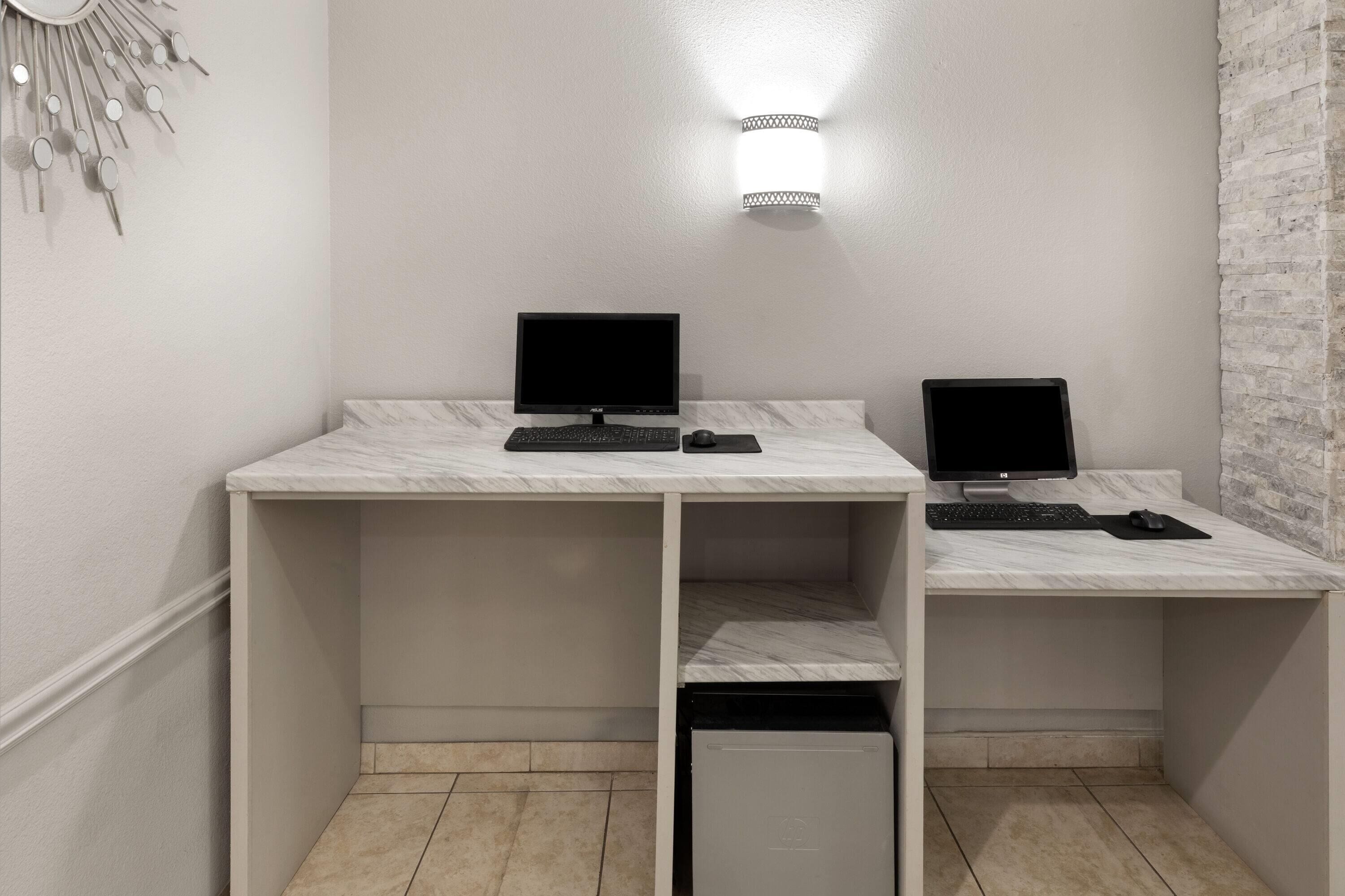 Um espaço de escritório moderno com duas mesas, cada uma com monitor de computador, teclado e rato. Há um aparelho de luz fixado na parede acima das mesas.