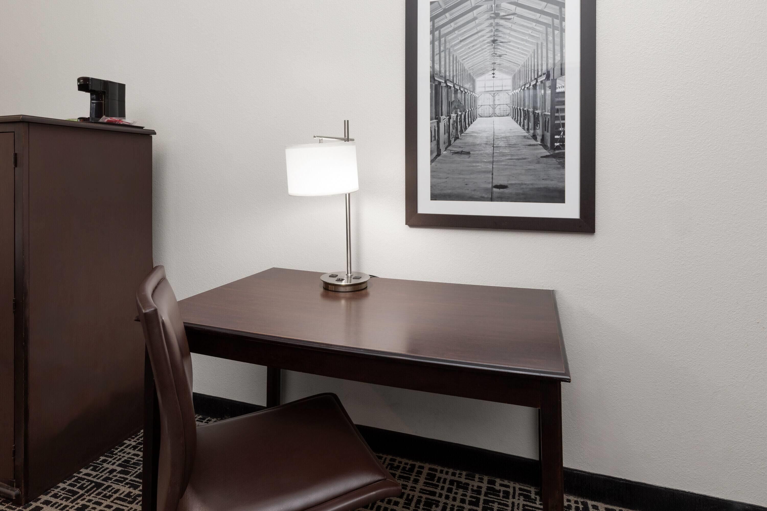 A hotel room with a wooden desk, a chair, a lamp, and a framed photograph on the wall.