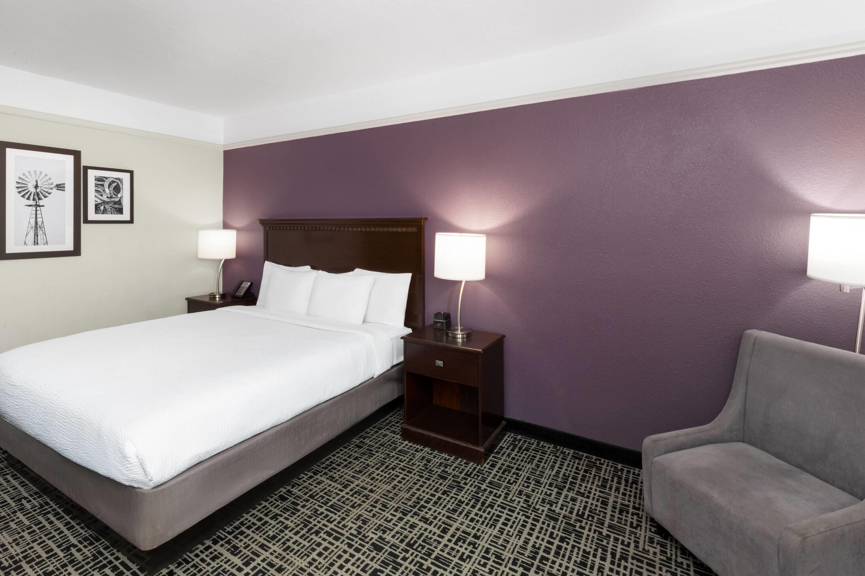 Habitación de hotel con una cama grande, dos mesitas de noche con lámparas, un sillón gris y alfombra estampada.