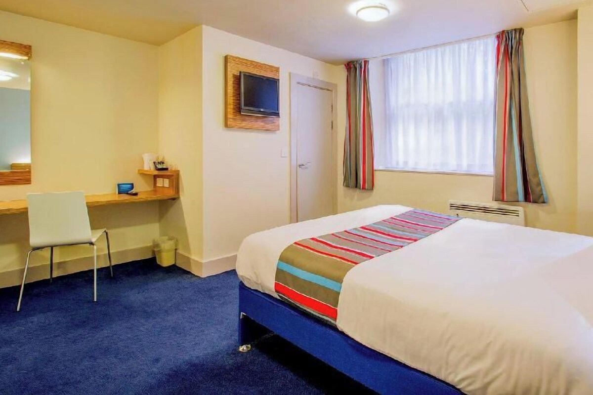 A hotel room with a bed, a desk, a chair, a television, and a window with curtains.