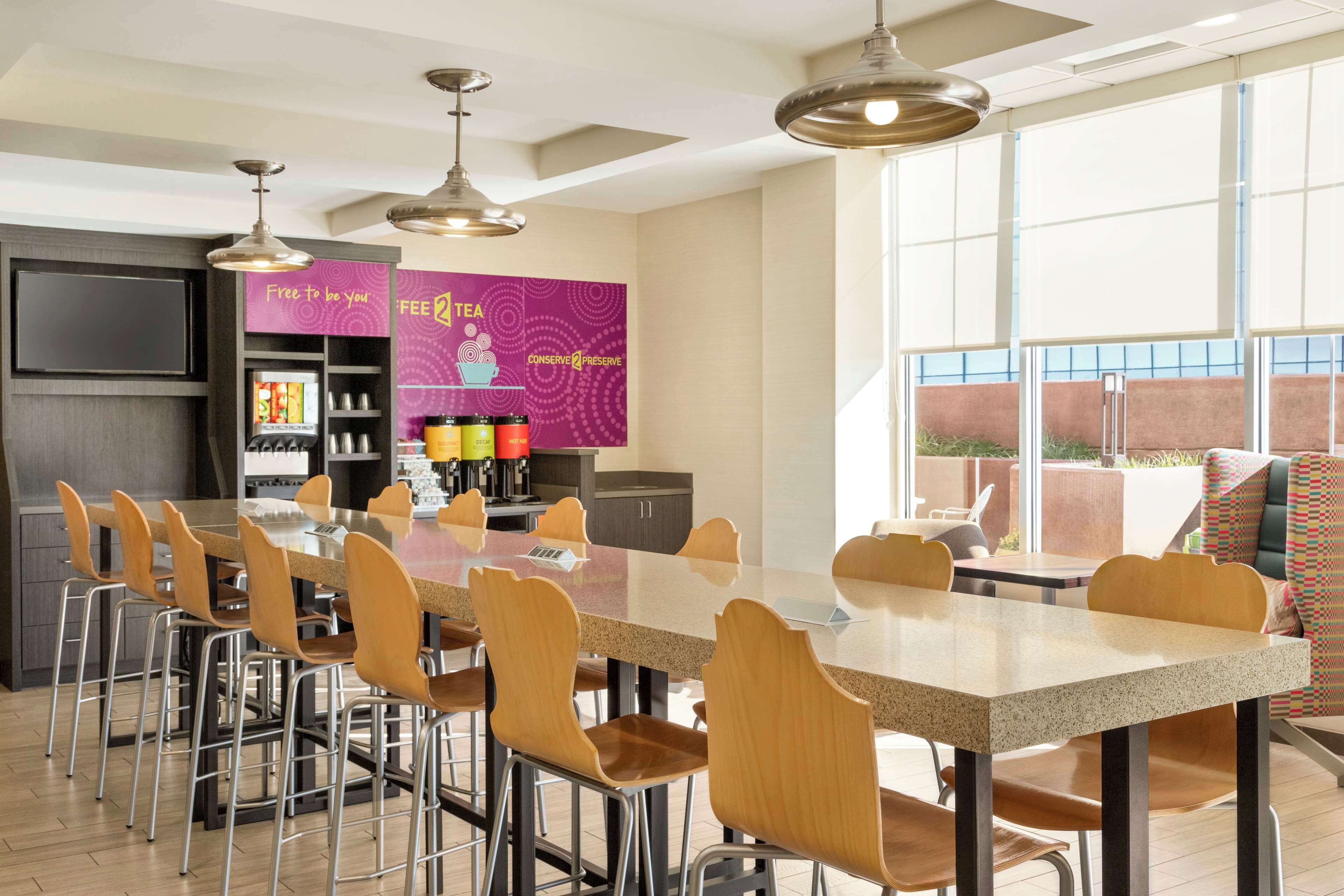 A modern dining area with a long table and chairs, a coffee station, and large windows offering a view.