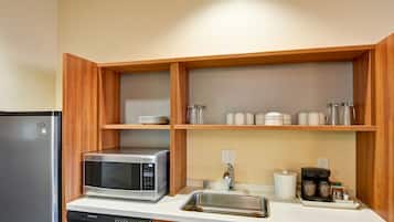 A compact kitchen with a stainless steel refrigerator, a microwave, and a dishwasher. There are wooden shelves above the countertop holding various kitchen items.