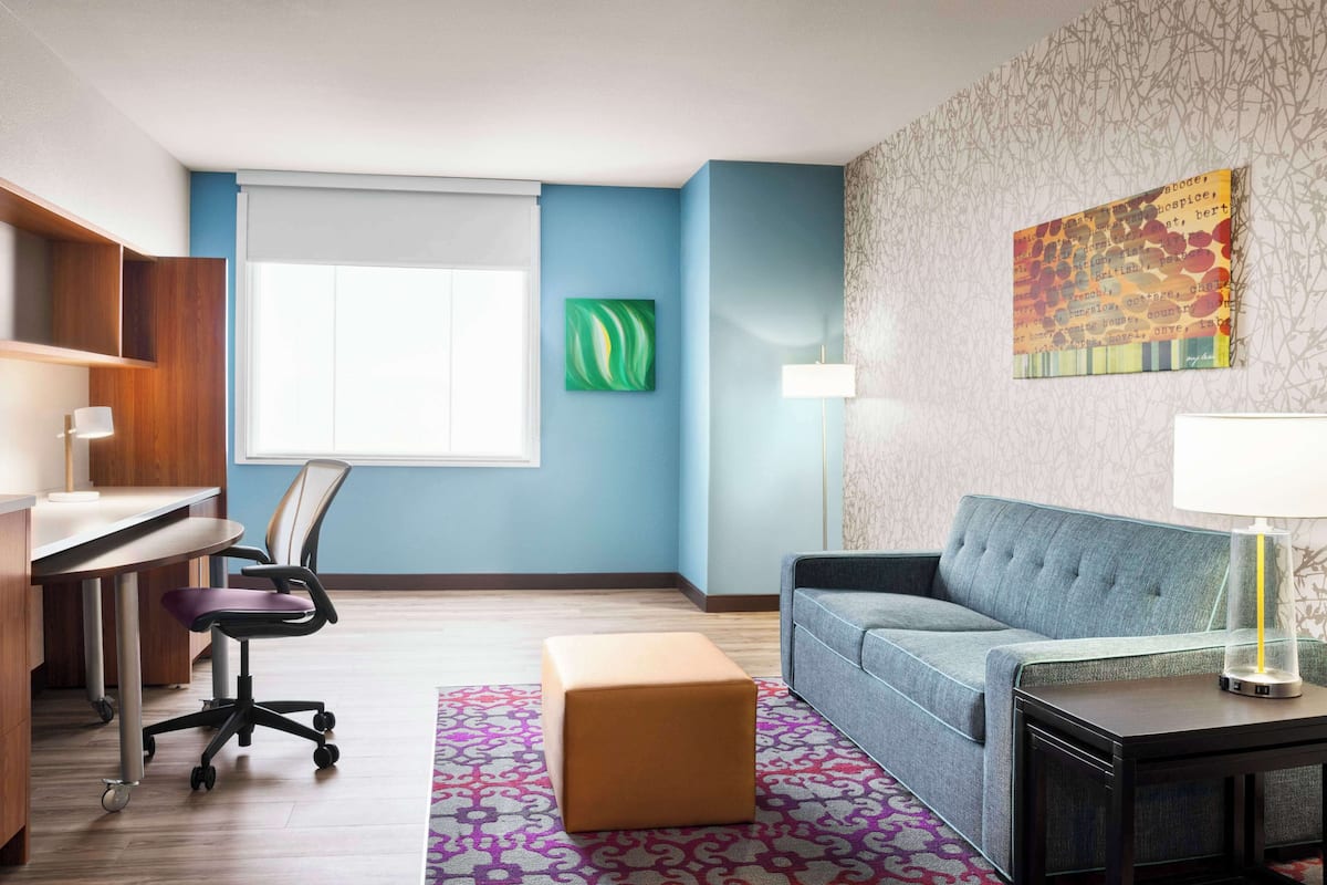 Ett modernt hotellrum med ett skrivbord, kontorsstol, soffa, pall och en lampa på ett sidobord.