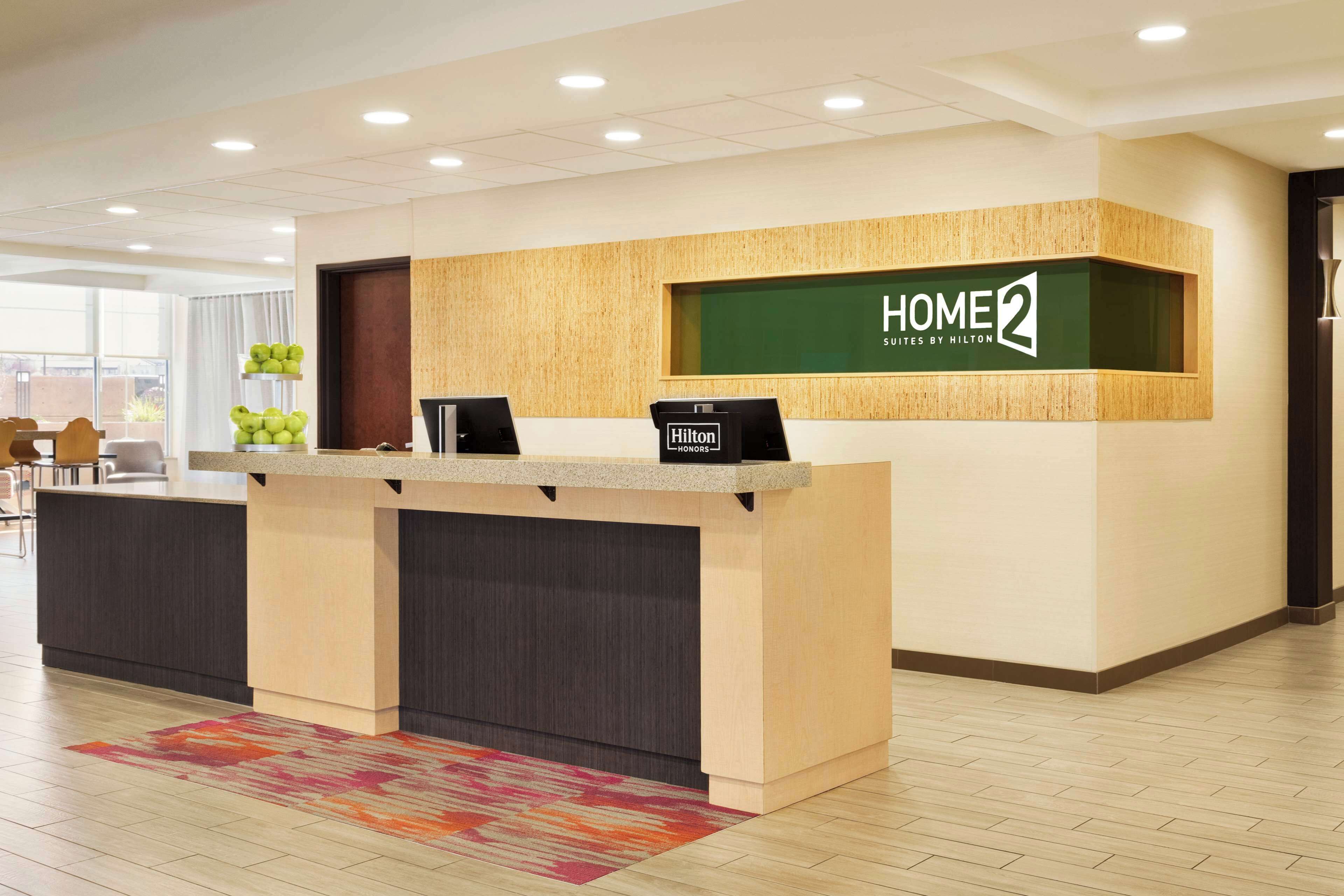 A hotel reception desk with a 'HOME 2' sign, a digital display, and a 'Hilton Honors' card reader.