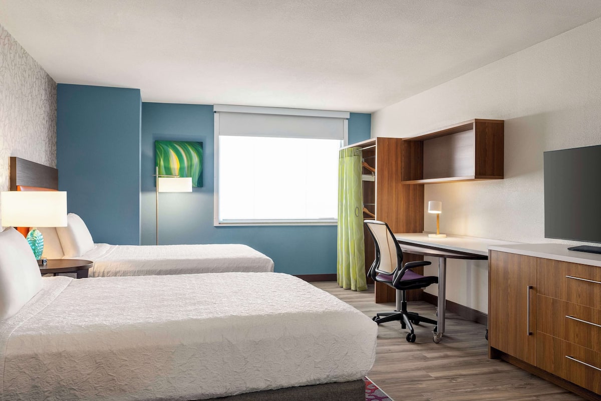 Ett hotellrum med två sängar, ett skrivbord, en stol, en TV och ett fönster med gardiner.