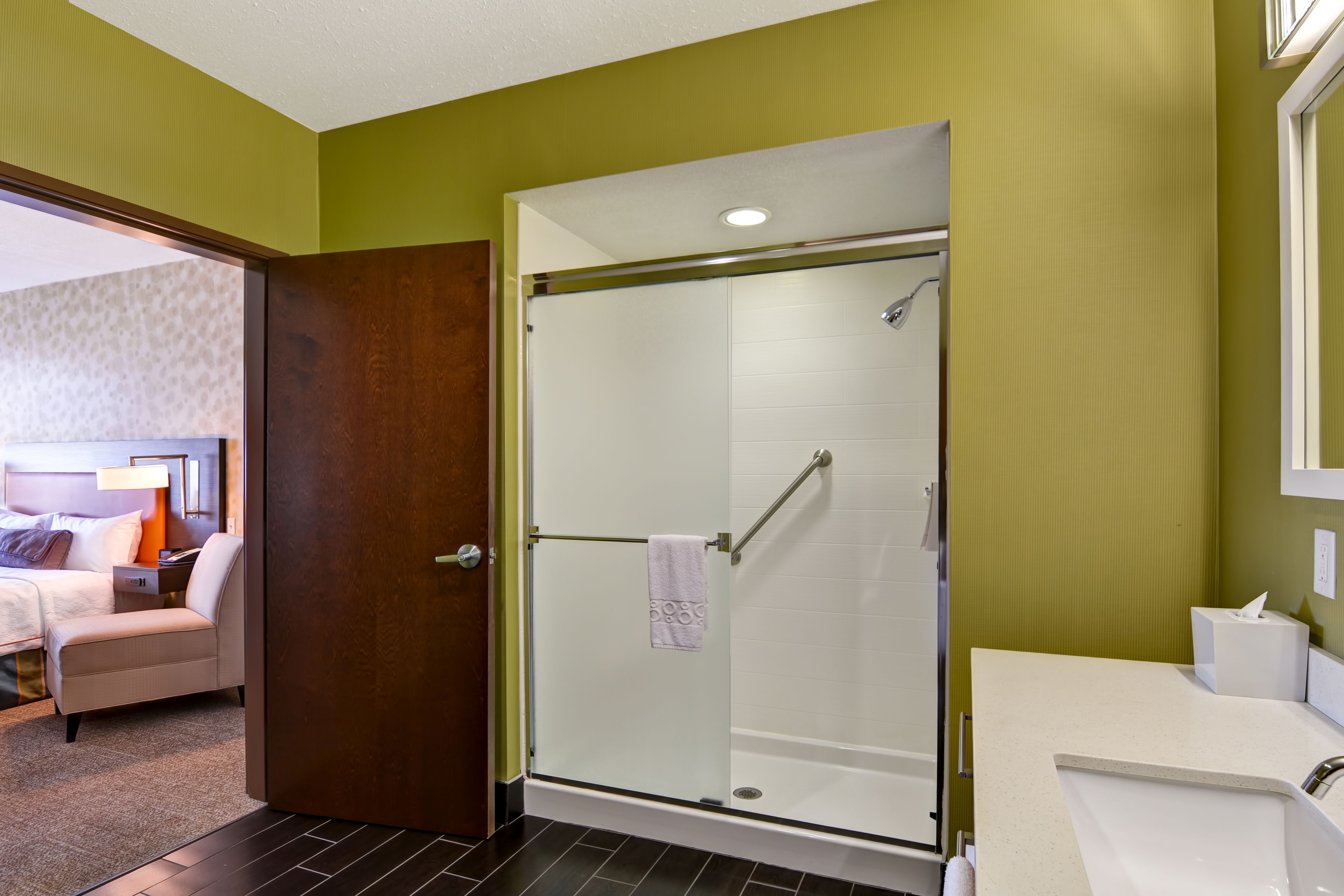 A hotel bathroom with a glass-enclosed shower, a white sink, and a bed in the adjacent room.