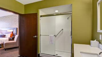 A hotel bathroom with a glass-enclosed shower, a white sink, and a bed in the adjacent room.