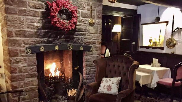 A cozy room with a stone fireplace, a red wreath, a plush armchair, a table with a menu, and a patterned carpet. - String of Horses (Brampton)