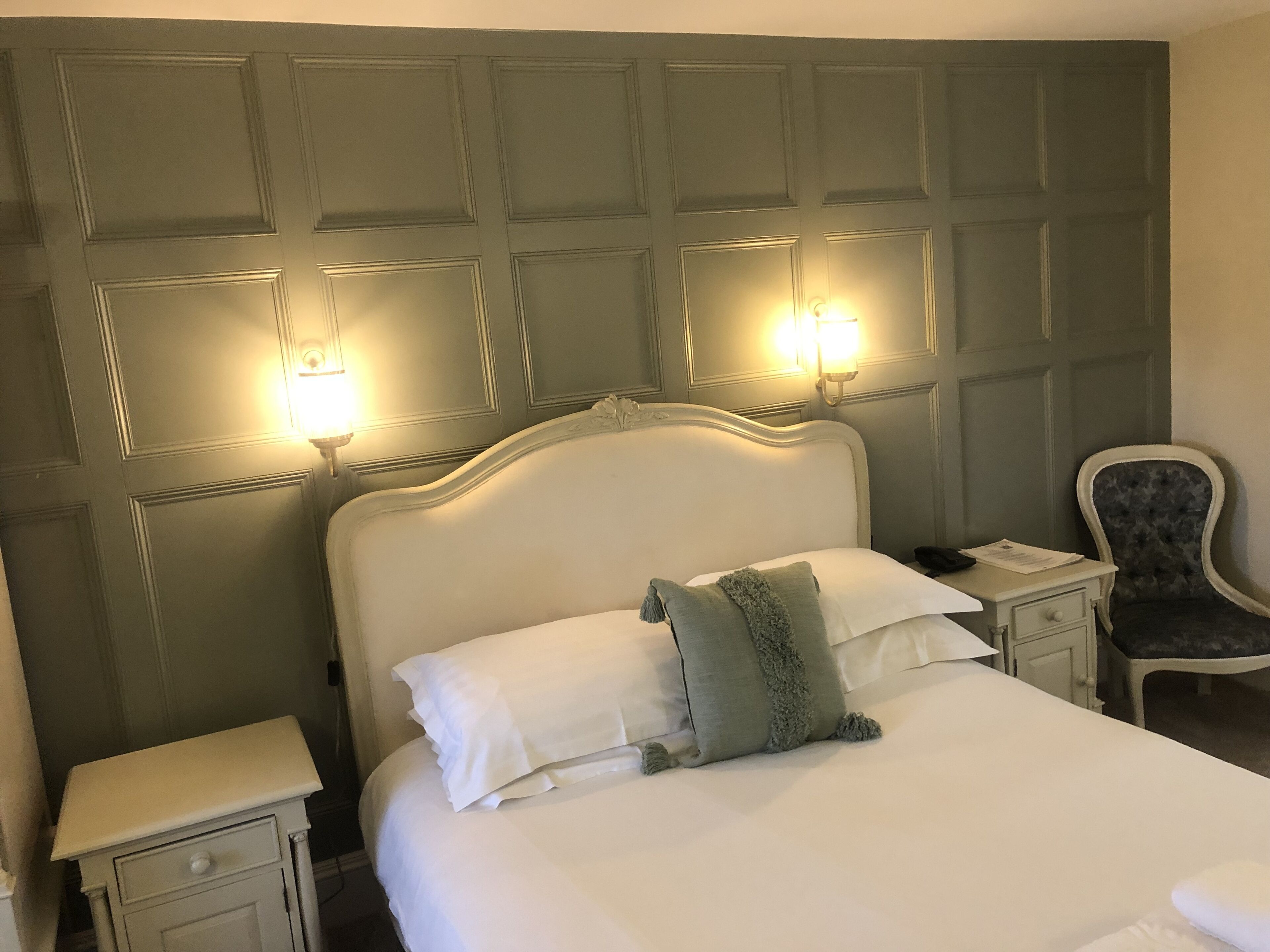 A neatly made bed with white linens and a green accent pillow, flanked by two bedside tables with lamps, in a room with a paneled headboard and a chair.