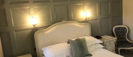 A neatly made bed with white linens and a green accent pillow, flanked by two bedside tables with lamps, in a room with a paneled headboard and a chair.