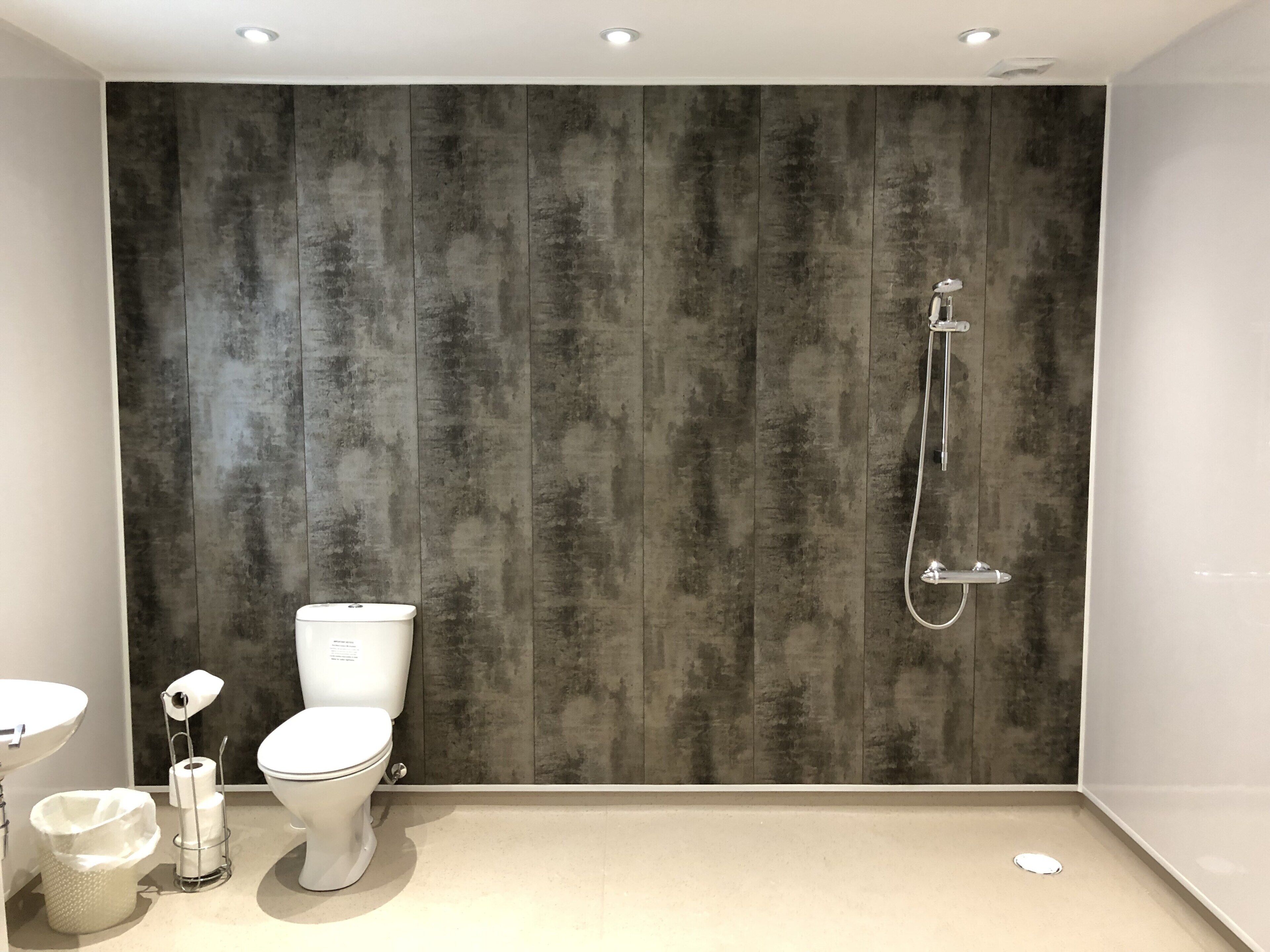 A modern bathroom with a grey wood-textured wall, a white toilet, a sink, and a shower with a handheld showerhead.