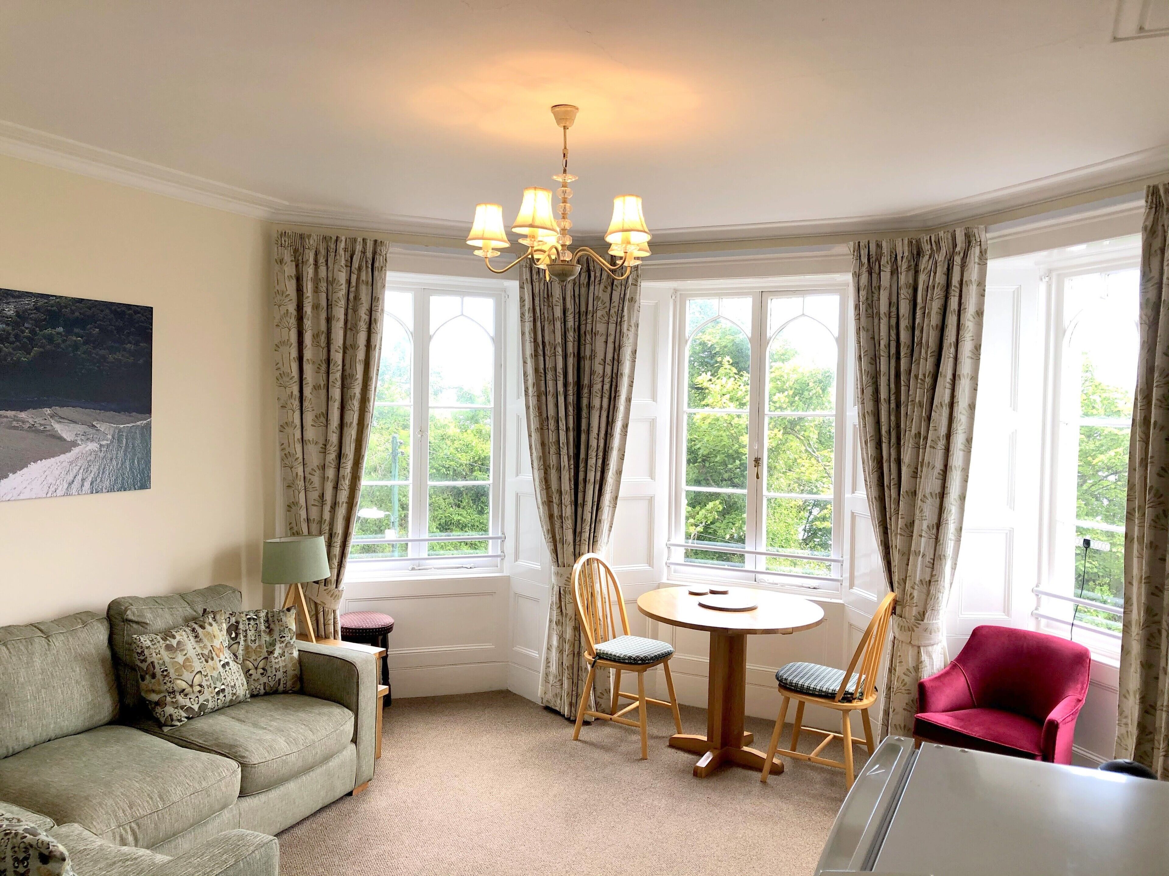 A room with a green sofa, a red chair, a round wooden table with chairs, a chandelier, and large windows with curtains.