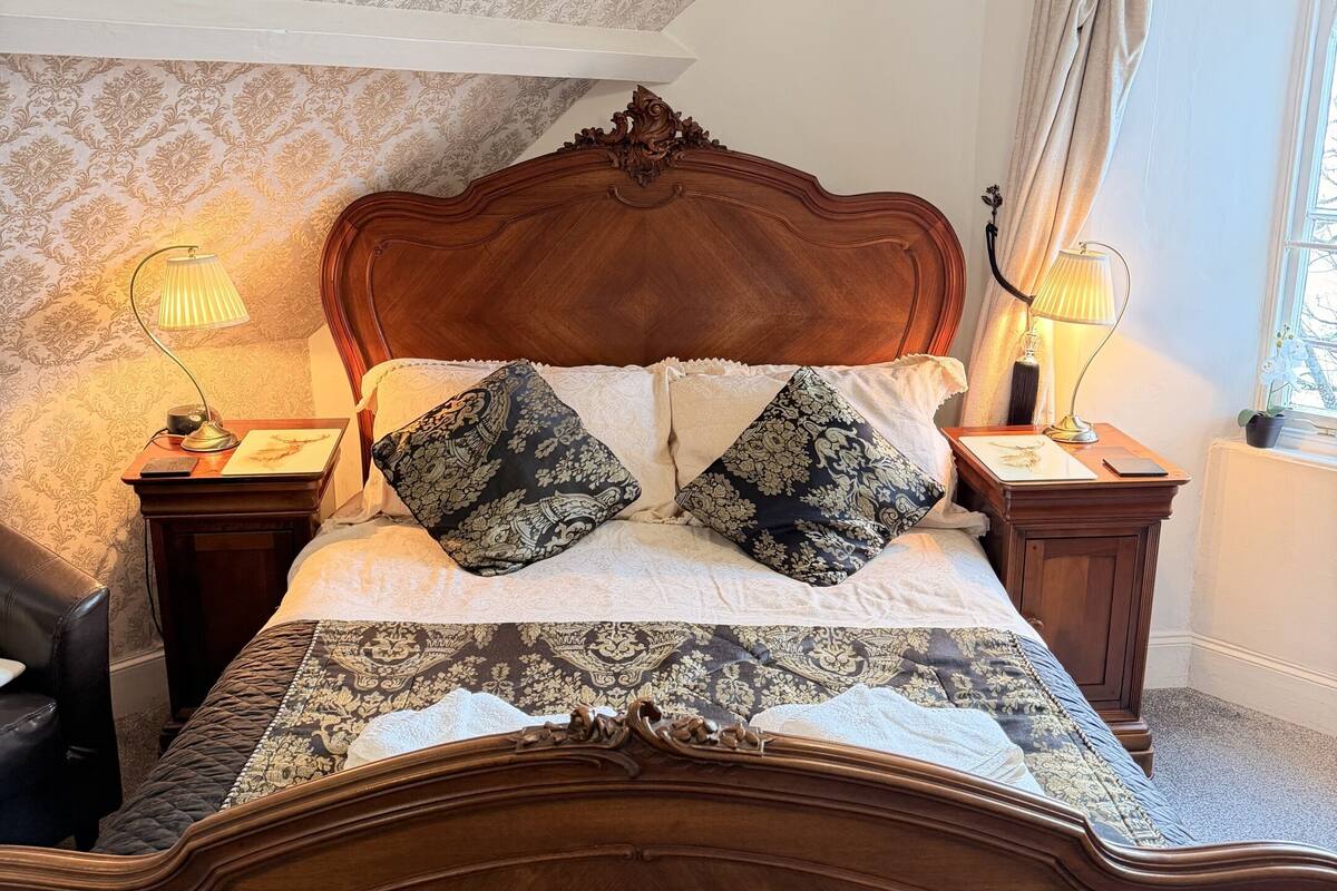 A bedroom with a wooden bed, patterned wallpaper, two bedside lamps, a window with curtains, and a chandelier.