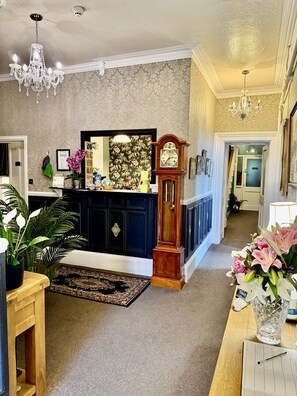 A spacious living room with a dark wooden cabinet, a large mirror, a grandfather clock, and a chandelier. - The North Cliff Hotel (Lynton)
