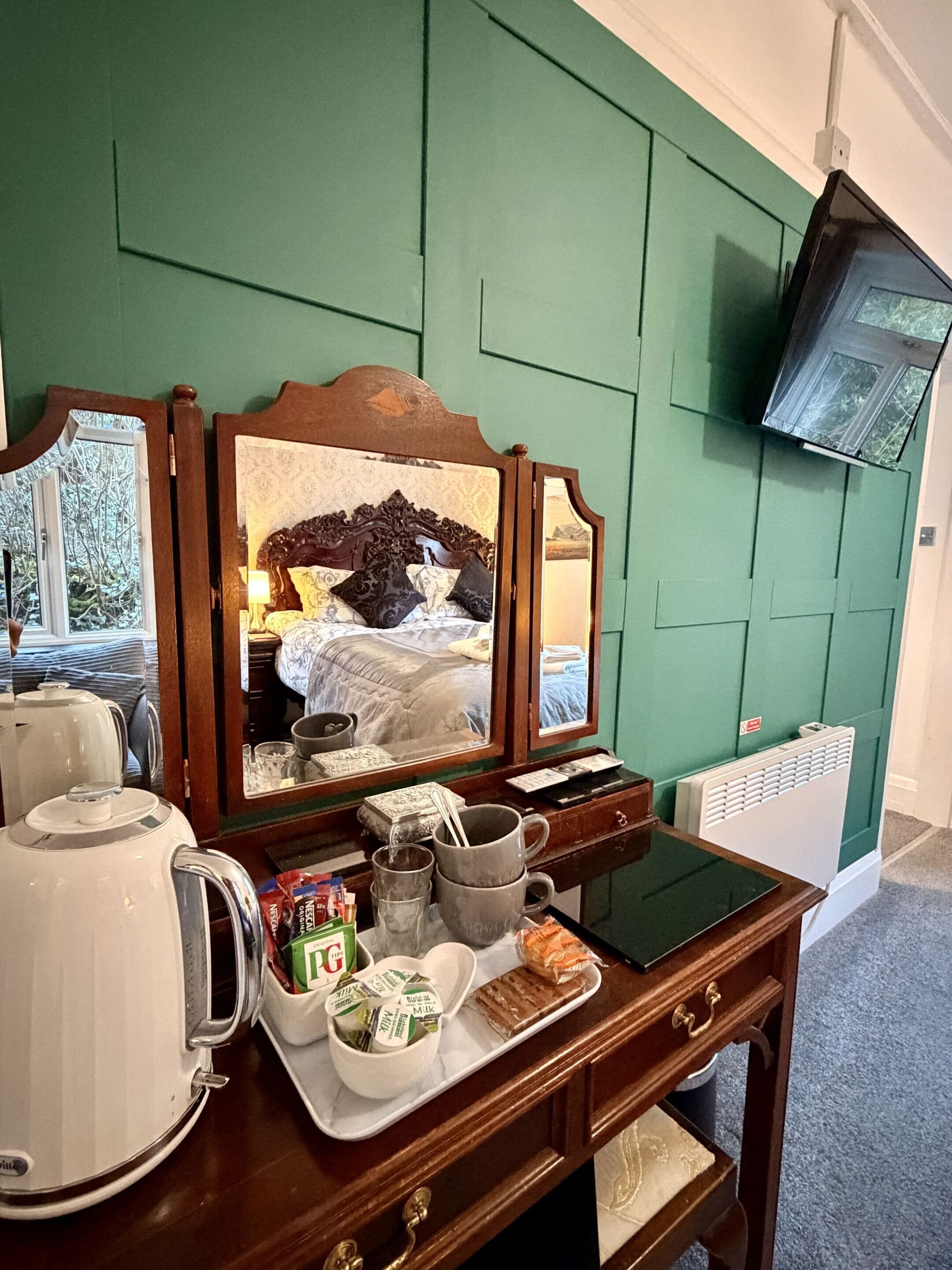 Ein Hotelzimmer mit einem Holzkoffer, einem Spiegel, einem an der Wand befestigten Fernseher und einem Fenster mit Blick auf Bäume.