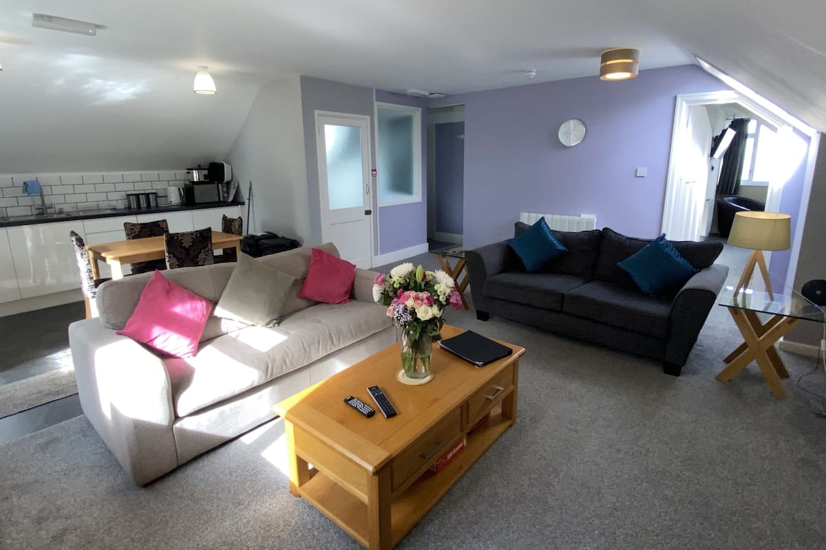 A living room with a sofa, a coffee table, and a lamp. There is a kitchen in the background with a window.