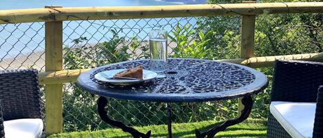 Mesa redonda con un plato y un vaso, rodeada de sillas de mimbre, sobre una zona cubierta de césped con vistas al mar y colinas.