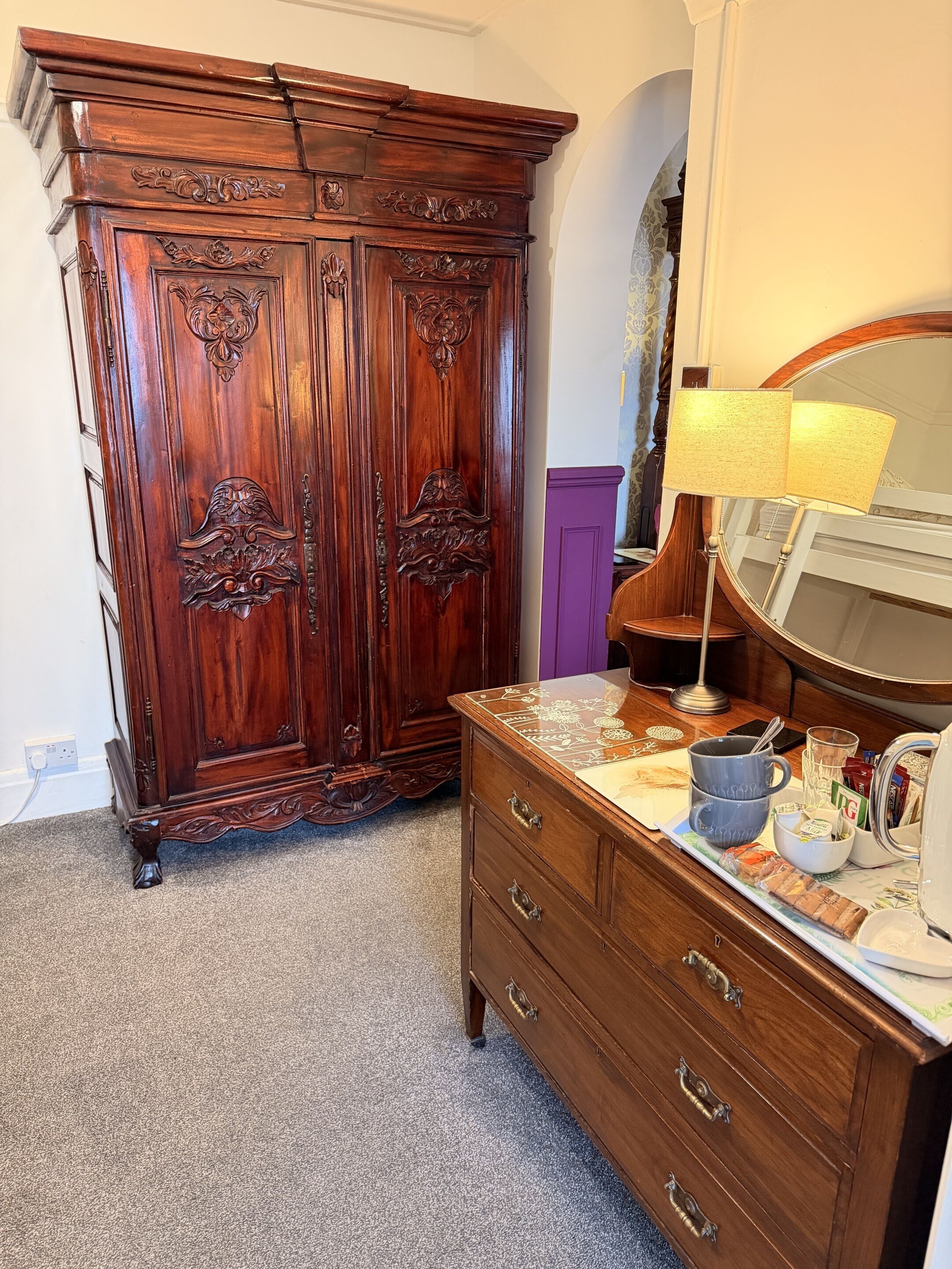 A wooden dresser with drawers and a mirror, a large wooden wardrobe with intricate carvings, and a lamp on a table.