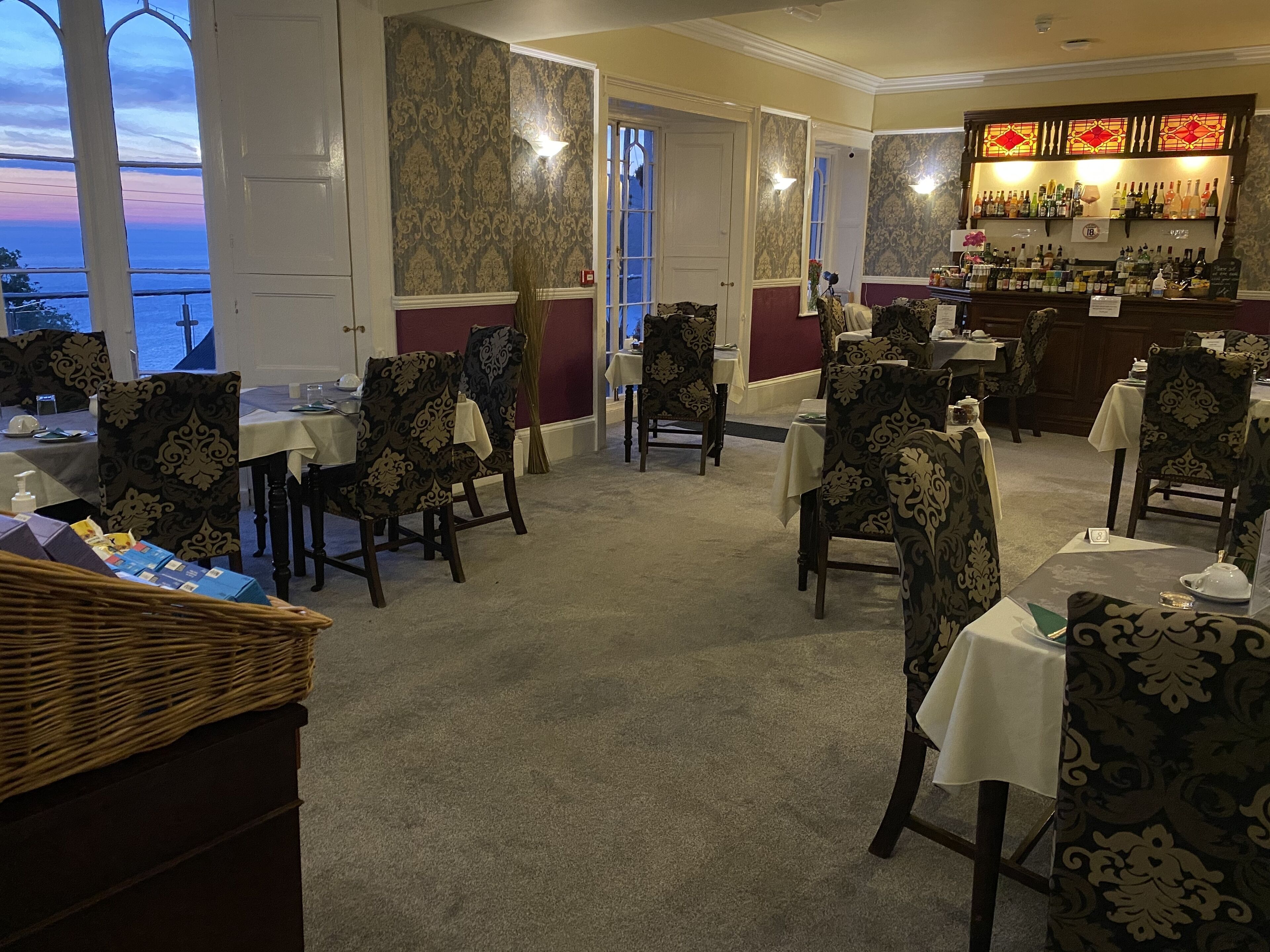 A dining area with patterned chairs and tables, a bar with various bottles, and a view of the sea through large windows.