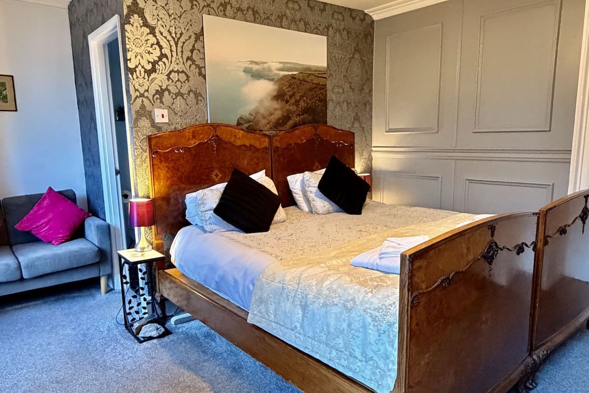 A bedroom with a wooden bed, a nightstand, a lamp, a sofa, and a large window with a view of the sea.