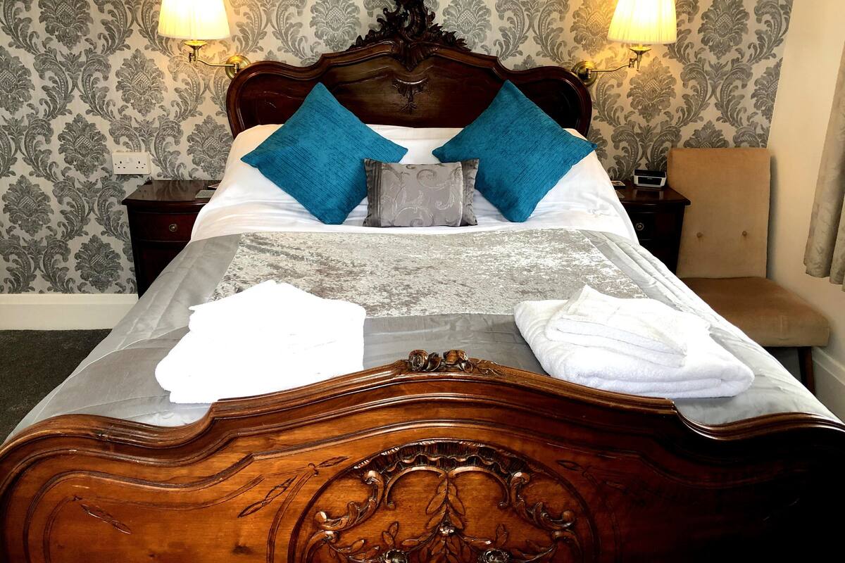 A neatly made bed with a wooden headboard, two blue pillows, and a grey throw. There are two bedside tables with lamps, a chair, and a wall with patterned wallpaper.