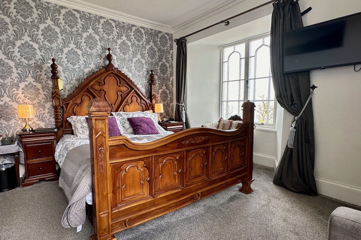 A bedroom with a wooden four-poster bed, a nightstand with a lamp, a window with curtains, and a flat-screen TV mounted on the wall.