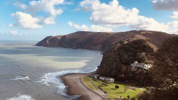 Un paisaje costero con una casa grande en una colina con vistas al mar, una playa rocosa y una zona verde con un sendero.