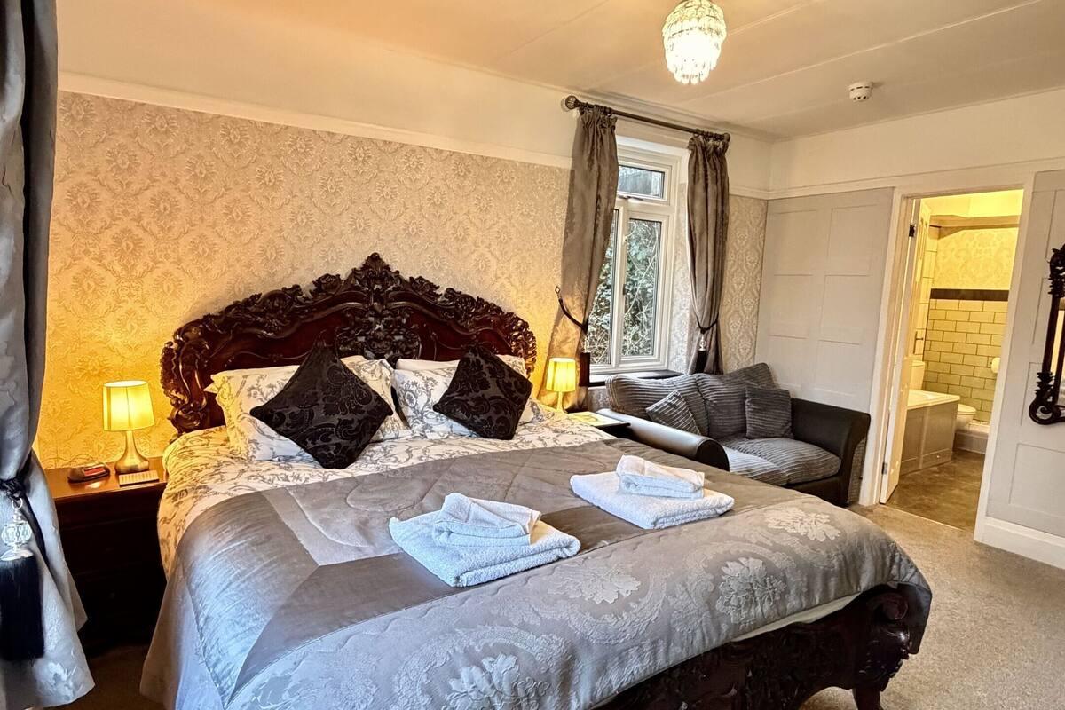 A bedroom with a large bed, a nightstand, a lamp, a window with curtains, and a view of the bathroom.