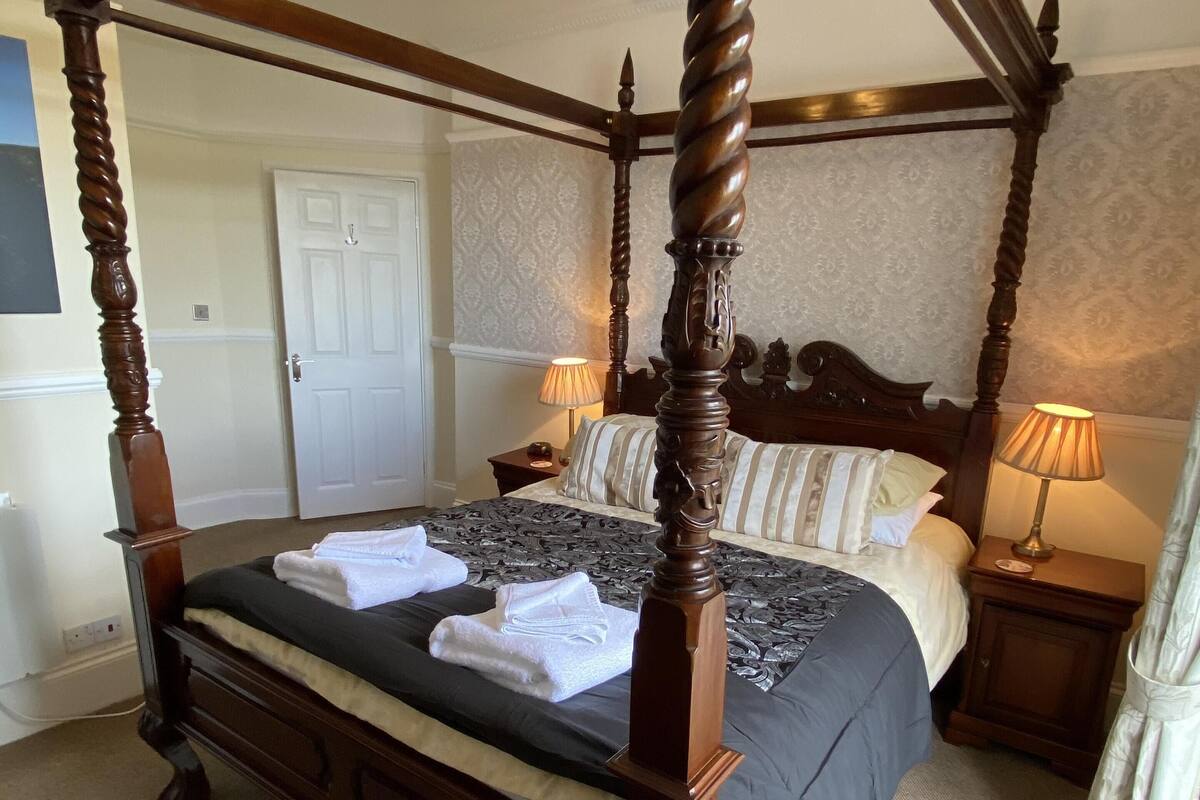 A four-poster bed with a dark wooden frame and a black bedspread, two white towels on the bed, and two bedside lamps on wooden nightstands.