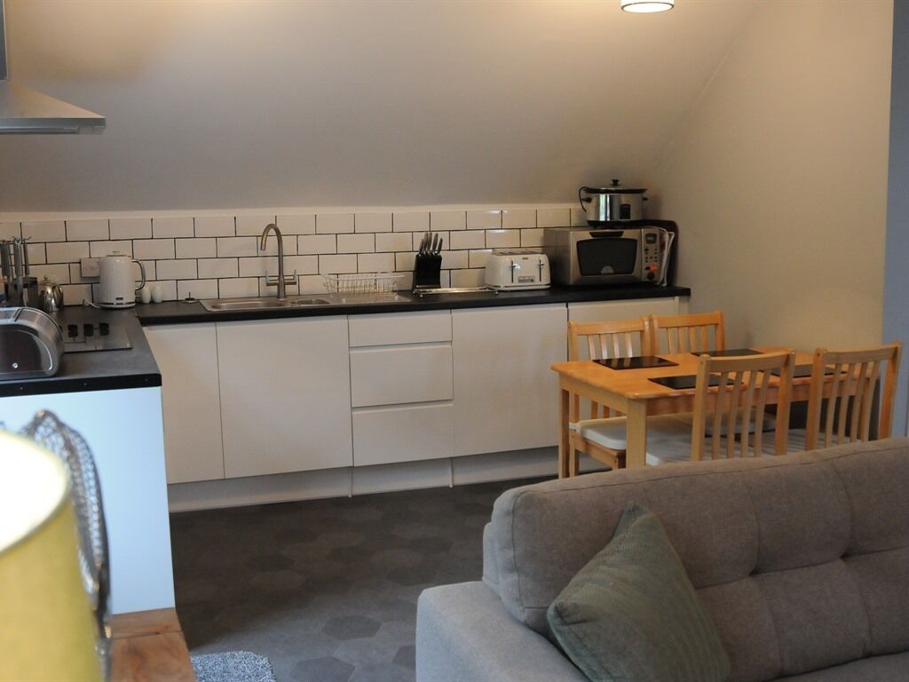 A compact kitchen with white cabinets, a black countertop, a toaster, a microwave, and a sink. There is a wooden dining table with chairs and a grey sofa in the living area.