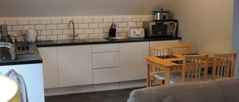 A compact kitchen with white cabinets, a black countertop, a toaster, a microwave, and a sink. There is a wooden dining table with chairs and a grey sofa in the living area.