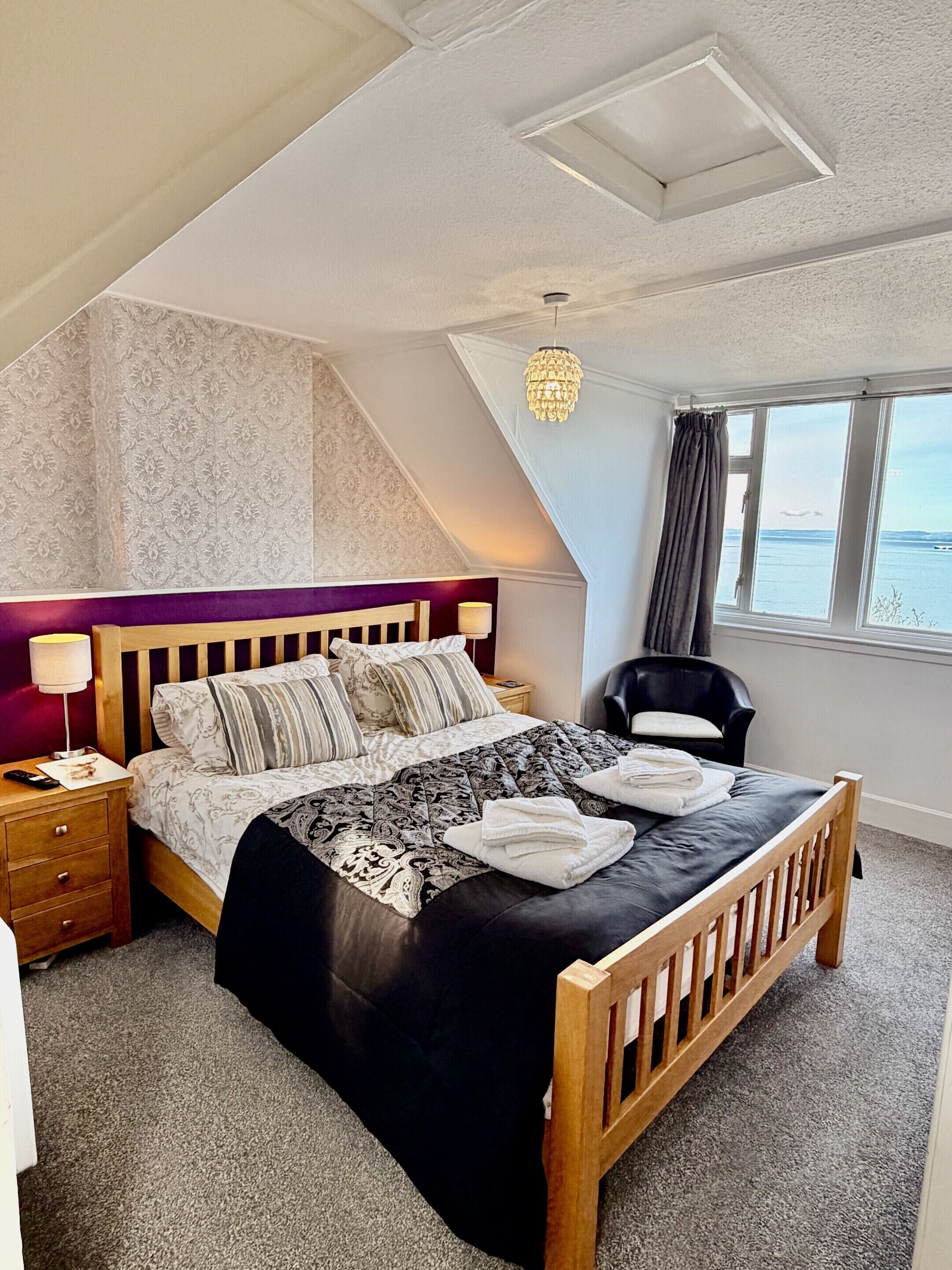 A bedroom with a bed, a nightstand, a lamp, a chair, a window with a view, and a chandelier.