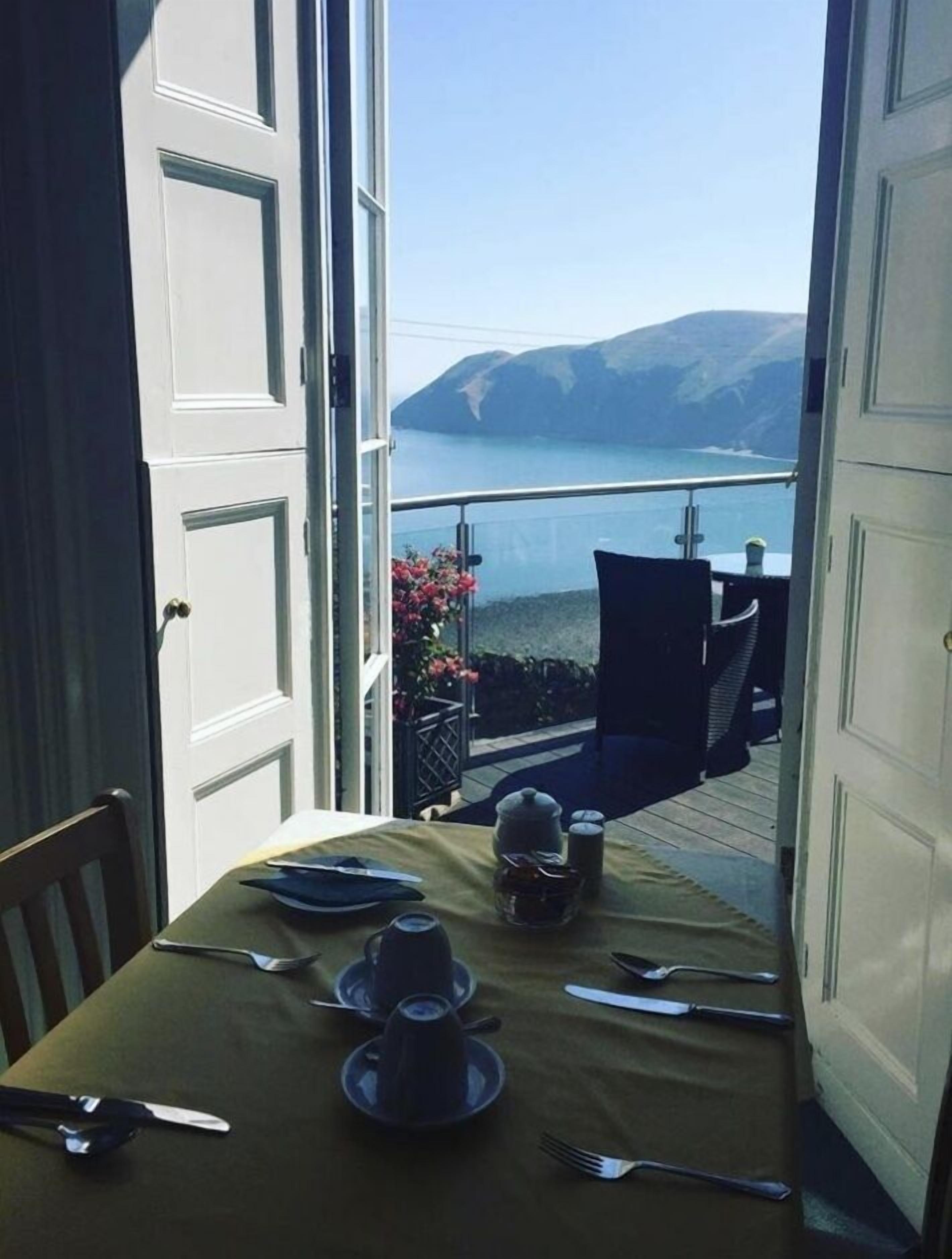 An open door leading to a balcony with a table set for two, overlooking a body of water and mountains.