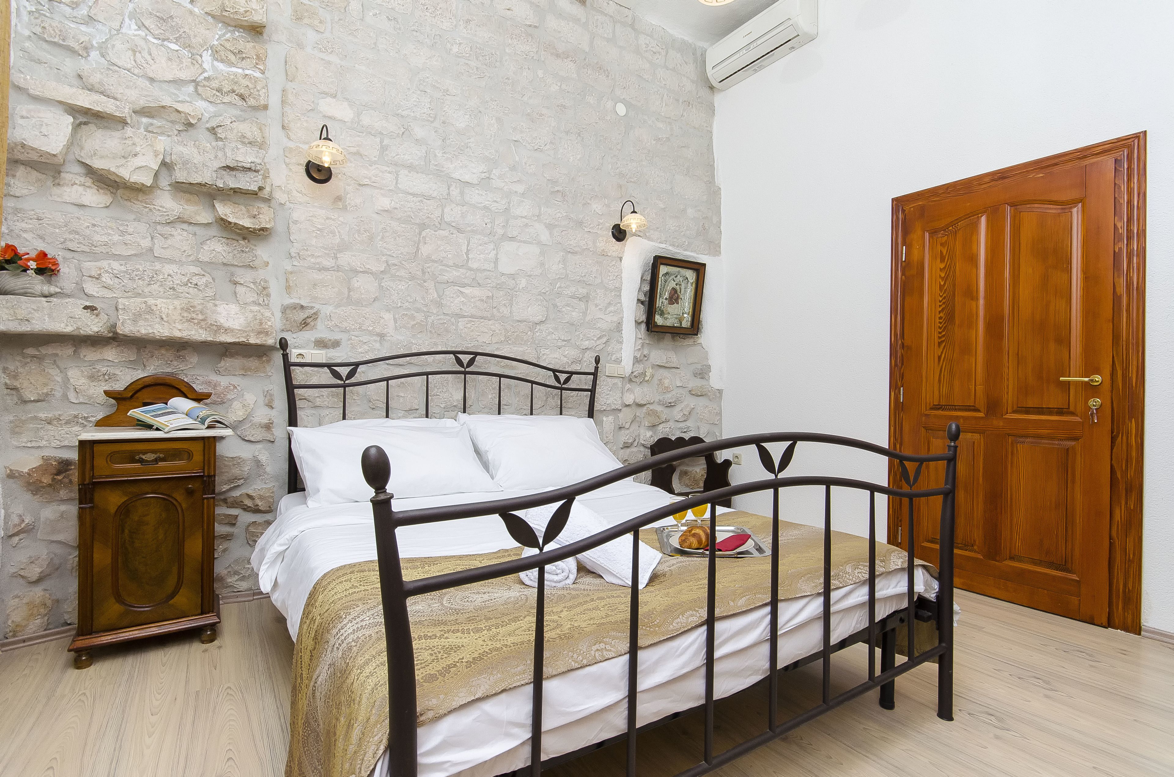 A bed with a metal frame, a wooden nightstand, a stone wall, and a wooden door.