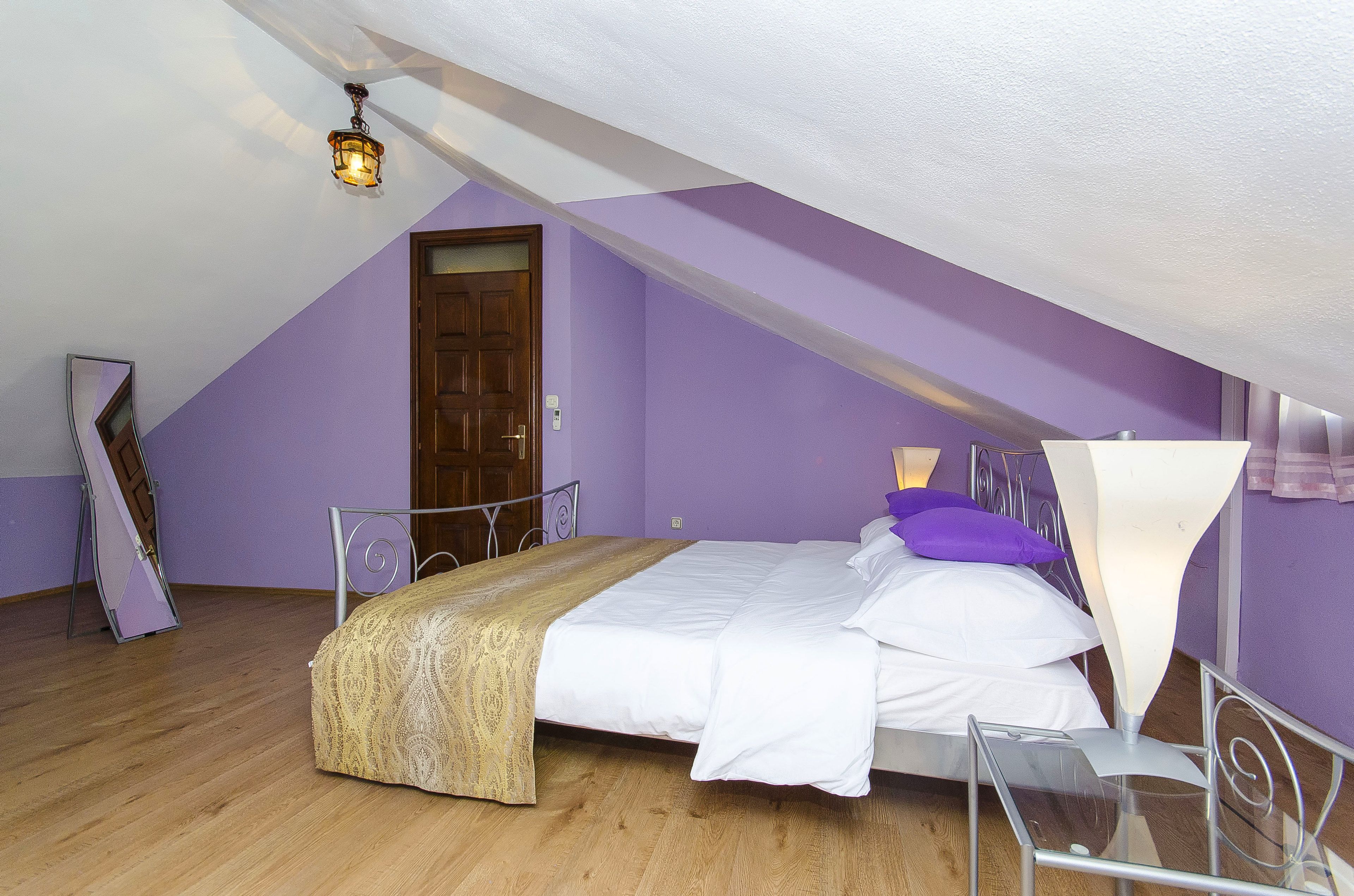 Ein Schlafzimmer mit schrägen Wänden, einem Bett mit weißer Bettwäsche, einer Holztür und einem kleinen Tisch mit Lampe.